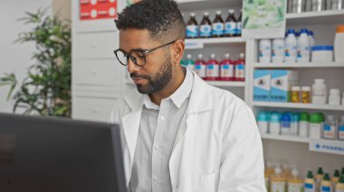 Beyaz önlüklü, odaklanmış bir Afro-Amerikan erkek eczacı modern bir eczane içinde bir bilgisayarda çalışıyor..