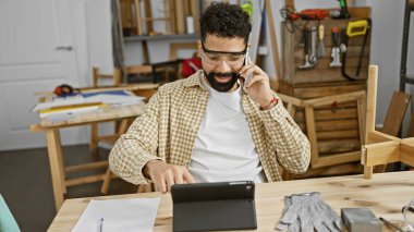 Marangozluk atölyesinde elinde aletler ve ahşap aletlerle konuşan kareli gömlekli sakallı bir adam..