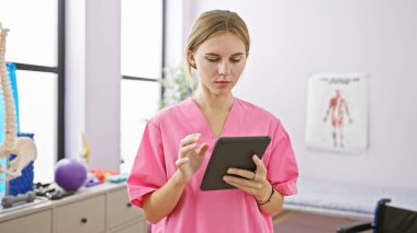 Pembe önlüklü sarışın kadın. Parlak bir rehabilitasyon kliniğinde elinde posterlerle tablet kullanıyor..