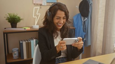 Kapalı alanda akıllı telefon kullanan gülümseyen kadın, ofis gündelik, kulaklık, eğlence, teknoloji, yetişkin, kadın, kitaplık, bitki, modern