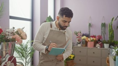 Önlük giymiş sakallı genç bir adam canlı buketler ve bitkilerle çevrili renkli bir çiçekçide notlar yazıyor..