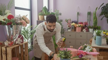 A young hispanic man with a beard works in a flower shop, using his phone amidst various plants and floral arrangements. clipart