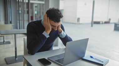 Stresli adam, modern işyerinde dizüstü bilgisayar ve akıllı telefonla baş ağrısı çekiyor..
