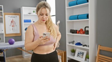 Rehabilitasyon kliniğinde akıllı telefon kullanan sarışın kadın, sağlık ve sağlık hizmetlerini betimliyor..