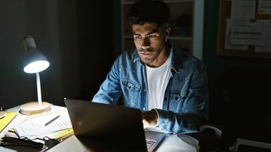 Laptop ve masa lambasıyla ofiste geç saatlere kadar çalışan, kendini işine adamış ve çalışkan İspanyol bir adam..