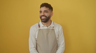 Önlük takan neşeli, genç, İspanyol bir adam saf sarı bir arka plana karşı duruyor, canlı ve cana yakın bir görünüm sergiliyor..
