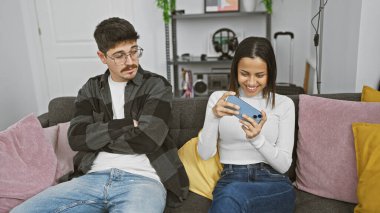 Sıkılmış bir adam rahat bir oturma odasında izlerken bir kadın telefonuyla oynuyor..