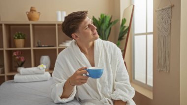 Yakışıklı genç adam sağlıklı bir spa 'da dinleniyor, beyaz bir bornoz giyiyor ve mavi bir fincan çay tutuyor, etrafı bitkilerle çevrili ve hafif ışıklandırmayla çevrili.