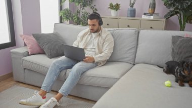 Dachshund 'un yanındaki koltukta kulaklıkla çalışan İspanyol bir adam modern bir apartman dairesinde oturuyor.