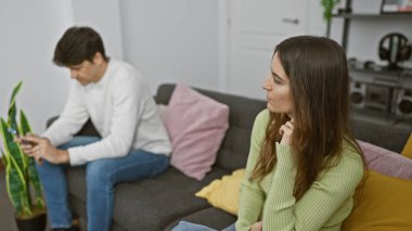 Düşünceli bir kadın ve erkek modern bir oturma odasındaki bir kanepede ayrı otururlar, gergin bir çiftin dinamiklerini yansıtırlar..