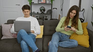 Gündelik giyinen bir kadın ve erkek, modern bir kanepede rahat bir oturma odasında otururken teknolojiyle meşgul olurlar..