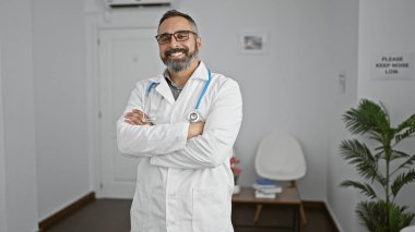 Gri sakallı, olgun İspanyol erkek doktor klinik koridorunda kollarını kavuşturarak profesyonellik ve deneyimi resmediyor..