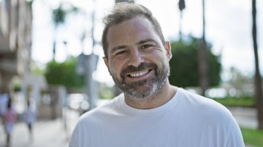 Yakışıklı, orta yaşlı, beyaz saçlı, şehir ortamında gülümseyen bir İspanyol..