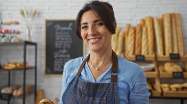 Fırında gülümseyen bir kadın, arka planda ekmek rafları ve karatahta menüsüyle, mavi gömlek ve önlük giymiş, orta yaşlı, misafirperver tavırlı.