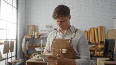 Genç adam pastanede bir deftere yazı yazarken, arka planda raflar ve taze ekmek ve pastalarla çevrili bir önlük giyiyordu..