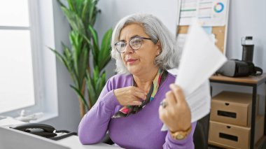 Mature woman fanning herself with paper in a modern office setting, expressing discomfort or heat. clipart