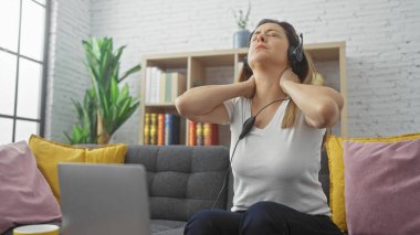 Boynu ağrıyan İspanyol kadın evde oturmuş kulaklık takıyor ve boynuna dokunuyor.