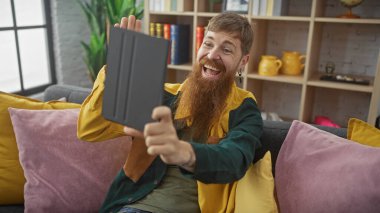Neşeli sakallı adam tabletle selfie çekiyor, parlak bir gülümseme, günlük kıyafetler, ev ortamı, rahatlık ve teknoloji gösteriyor..