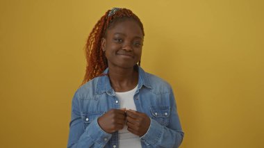 Kot ceketli neşeli, Afro-Amerikalı bir kadın sarı bir arka plana karşı poz veriyor, kendine güvenen bir gülümsemeyle..