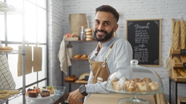 Yakışıklı, sakallı, fırında gülümseyen, arka planda ekmek ve hamur işleri olan bir İspanyol.