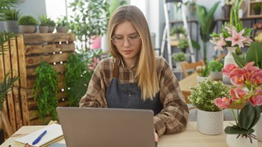 Genç bir kadın, canlı bir çiçekçide çiçeklerin arasında dizüstü bilgisayarda çalışıyor. Girişimcilik ve çiçekçiliğin vücut bulmuş hâli..