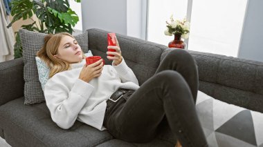 Rahat bir kadın, rahat bir oturma odasındaki kanepede akıllı telefonuyla fincanın keyfini çıkarır..