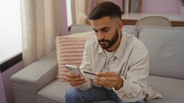 Oturma odasındaki koltukta oturan sakallı genç İspanyol bir adam akıllı telefon ve kredi kartı kullanıyor ve online bir işleme konsantre oluyor.