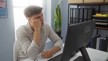 Ofisteki genç adam, çeşitli ofis malzemeleri ve raflarıyla çevrili bir bilgisayar monitörünün önünde otururken düşünceli görünüyor..