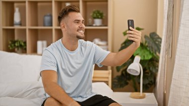 Gülümseyen genç bir adam rahat bir yatak odası ortamında modern dekorla selfie çekiyor, rahat bir yaşam tarzı yayıyor..