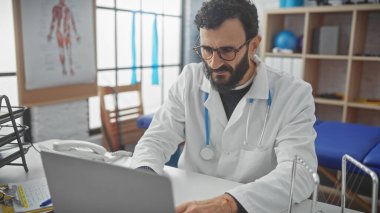Klinikte dizüstü bilgisayarda çalışan sakallı doktor. Arka planda anatomi posteri var.