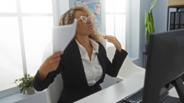 Ofiste çalışan bir kadın profesyonel bir ortamda bilgisayarın önünde otururken kendini evraklarla yelpazeliyor.