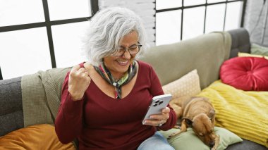 Neşeli, gri saçlı bir kadın akıllı telefonuyla köpeğiyle rahat, kapalı bir ortamda etkileşim kurmaktan hoşlanır..