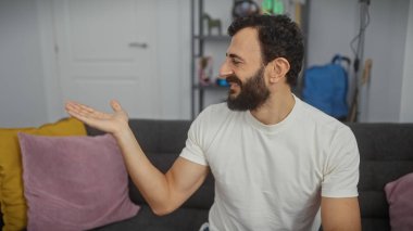 Beyaz tişörtlü sakallı bir adam modern bir oturma odasında uzanmış bir elle gülümsüyor ve jestler yapıyor.