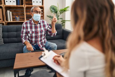 Tıbbi maskeli neşeli kıdemli adam, psikoloji kliniğinde otururken neşeli bir şekilde kenarı işaret ediyor. Covid 'in ortasında güven ve iyimserlik yayıyor.