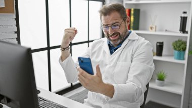 Beyaz laboratuvar önlüğü giymiş, orta yaşlı, neşeli, beyaz saçlı İspanyol bir adam, parlak, modern bir klinikte akıllı bir telefona bakarken yumruğuyla kutlama yapıyor..