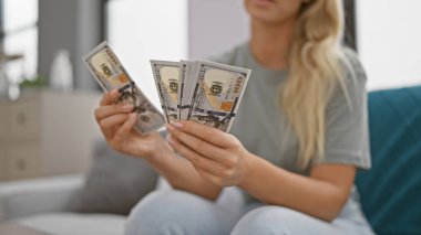 Genç sarışın bir kadın modern bir oturma odasında bize dolar sayıyor, finansal konuları resmediyor..