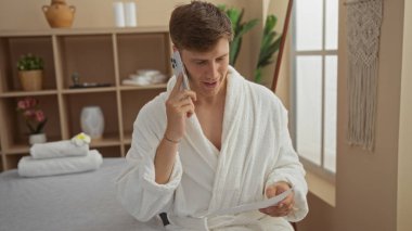 Beyaz cüppeli genç adam, elinde bir kaplıcada kağıt tutarken telefonla konuşuyor. Etrafı bitkiler ve dekorlarla dolu.