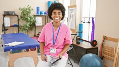 A smiling african american woman healthcare professional seated in a rehab clinic room, displaying confidence and approachability. clipart