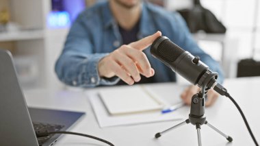 Bir adam mikrofon ve dizüstü bilgisayarla modern bir stüdyoda podcast yaparken el kol hareketi yapar.