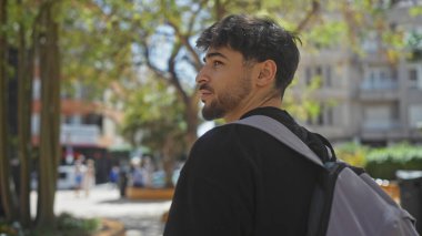Sırt çantalı yakışıklı sakallı adam canlı bir şehir parkına bakıyor..