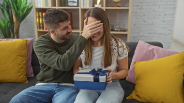 A man surprises a woman with a gift in their cozy living room, expressing love and togetherness. clipart