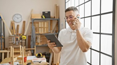 Marangozluk atölyesinde tablet ve telefon kullanan gülümseyen İspanyol adam.