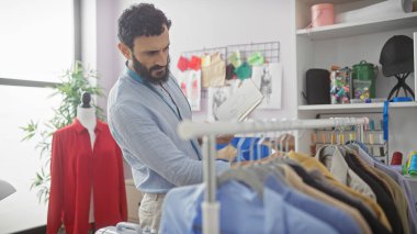 Sakallı terzi, iyi aydınlatılmış bir moda stüdyosunun tasarımlarını inceliyor..