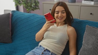 Genç bir İspanyol kadın oturma odasında elinde kırmızı bir telefonla mavi bir koltukta oturan bir sesli mesaj gönderiyor..