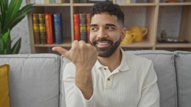 Yakışıklı İspanyol bir adam başparmağıyla evi işaret ediyor, modern bir dairede kitap rafı ve kanepesi var..