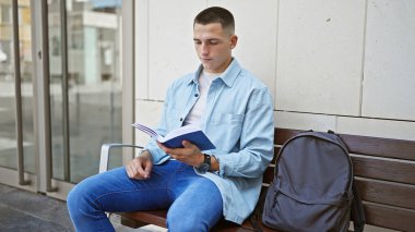 A young man in casual attire reading outdoors on a university campus bench, denoting a studious atmosphere. clipart