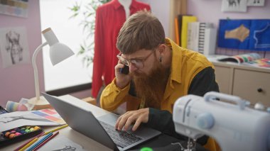 Sakallı adam, terzi atölyesinde dizüstü bilgisayar ve telefon kullanıyor. Dikiş malzemeleri ve renkli kumaşlarla.