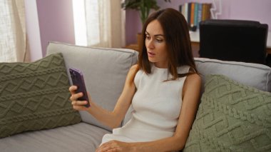 Evde yeşil yastıklı akıllı telefon kullanan genç bir kadın, şaşırmış ya da odaklanmış görünüyor, basit ama anlamlı bir iç mekan sahnesi sunuyor..