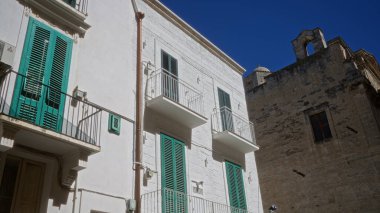 Puglia 'daki eski kasaba binalarının sokak manzarası, açık mavi gökyüzünün altında karakteristik yeşil panjurları ve balkonları var..