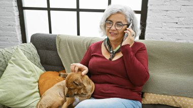 Gri saçlı bir kadın evdeki kanepede dinleniyor, evde telefonla konuşurken köpeğini okşuyor..